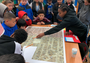 Students stand around a table with a large map on it. One student is pointing to a location on the map with a pencil.