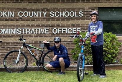cyclists stand with their bikes in front of a building with signage that reads “Yakin County Schools Administrative Offices” 